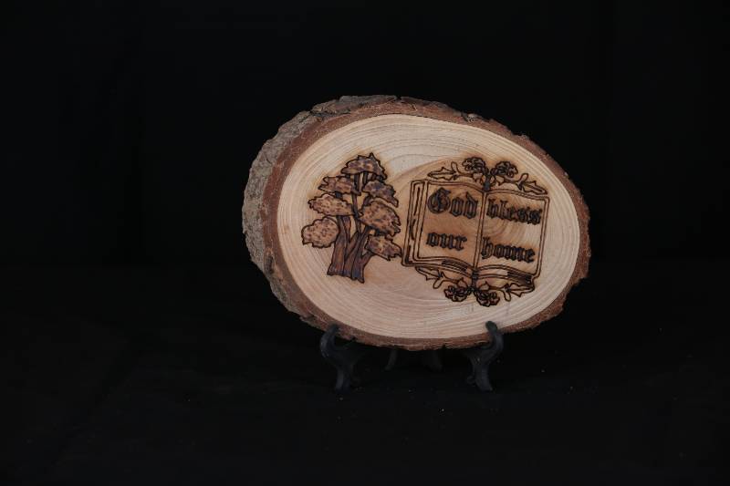 Handcrafted Wood Slice Plaque – “God Bless Our Home” with Book and Cedar Tree Design
 لوحة خشبية منقوشة يدويًا –  بتصميم كتاب وشجرة ارز